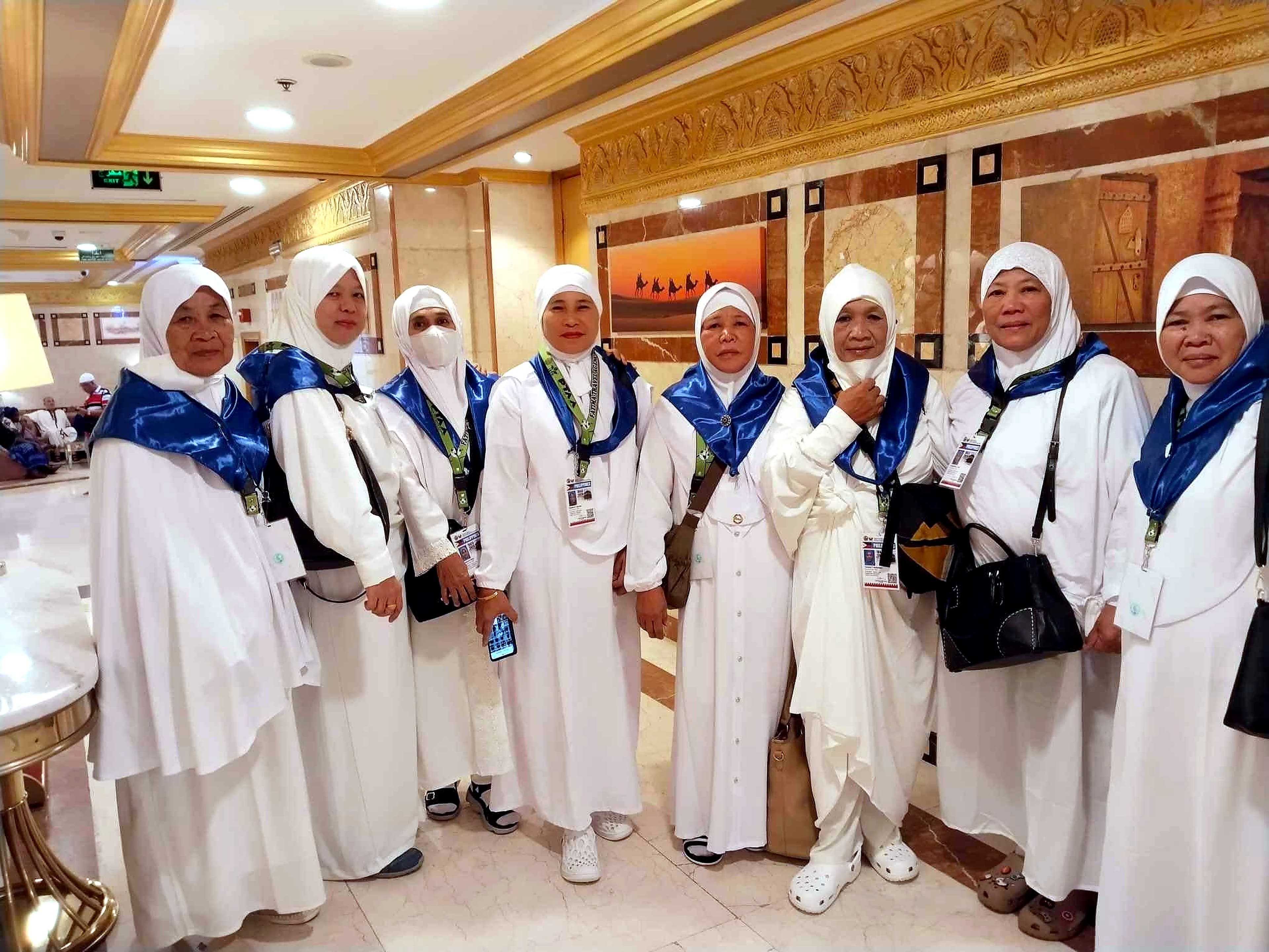 Eight women pilgrims in white garments and blue sashes posing together in an ornate hotel lobby.