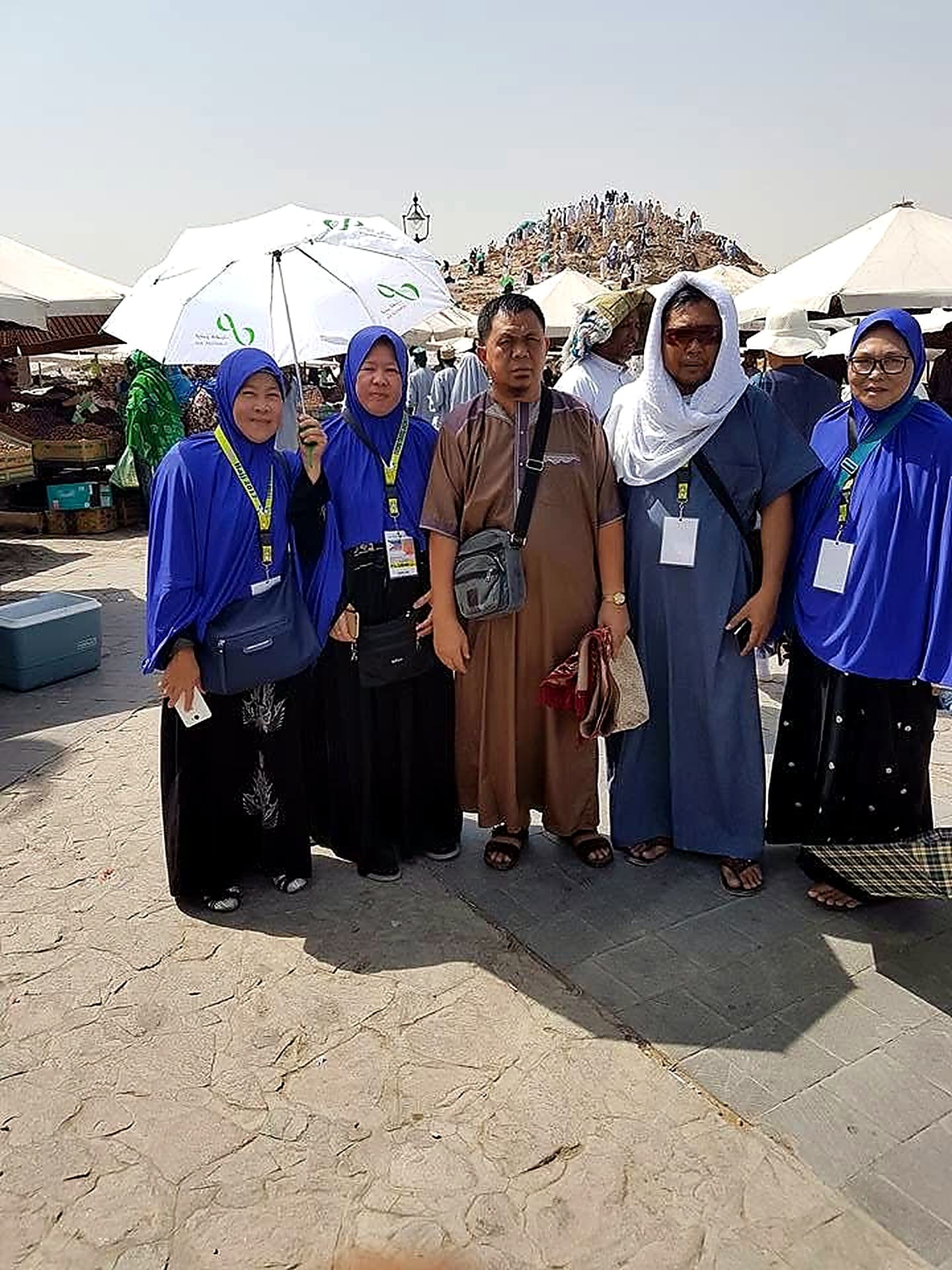Five pilgrims standing together in bright sunlight, with a hill crowded with pilgrims behind them.
